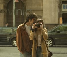 a man and woman standing next to each other on the street taking pictures with their cell phones