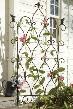 an iron trellis with pink flowers growing on it's sides and a watering can in the foreground