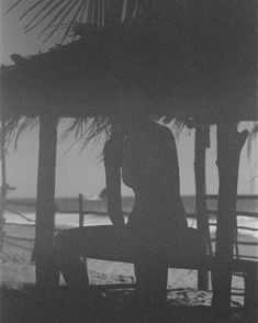 a man sitting on top of a bench under a thatched roof next to the ocean