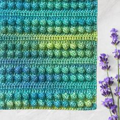 a crocheted square with blue and green stripes next to purple flowers on a white surface