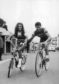 two people riding bikes on a street with other people standing around the building behind them