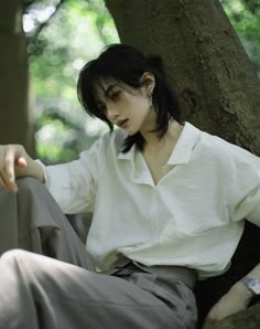 a woman sitting on the ground in front of a tree wearing a white shirt and gray pants