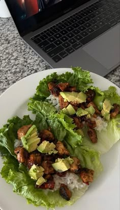 a white plate topped with lettuce covered in meat and avocado next to a laptop computer