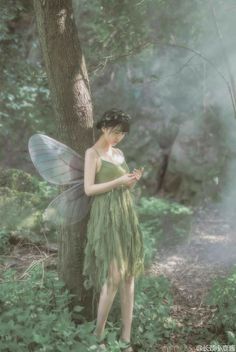 a woman dressed as a fairy standing next to a tree in the forest holding a cell phone