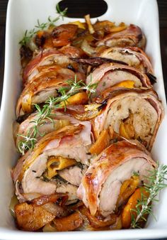 meat and vegetables in a white casserole dish on a wooden table with a sprig of rosemary