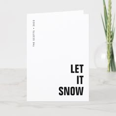 a white card with the words let it snow printed on it next to a plant