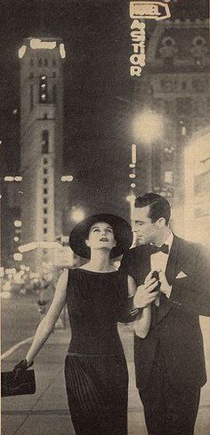 an old black and white photo of a man and woman walking down the street at night