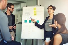 three people standing in front of a white board with sticky notes on it and pointing to them