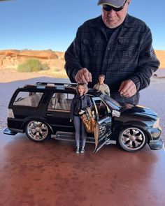 a man standing next to a toy car with a doll in the front and another person behind it