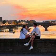 a man and woman sitting on a wall next to the water at sunset, with a bridge in the background