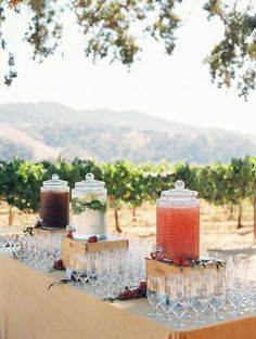 three jars filled with liquid sitting on top of a table covered in wine glasses and strawberries