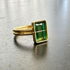 You won't be able to take your eyes off of this radiant cut green tourmaline ring. Aptly named, this stone radiates an array vibrant green hues. The double band compliments the crossbar and provides a luxurious contrast to the stone's geometric and longitudinal facets. A true one of a kind. 18k gold. 2.42 carat radiant cut green tourmaline. Size 7. Green Tourmaline Ring For May Birthstone, Modern Tourmaline Emerald Cut Rings, Modern Tourmaline Rings With Emerald Cut, Modern Tourmaline Rings In Emerald Cut, Fine Jewelry Green Rings With Rectangular Stone, Modern Green Emerald-cut Jewelry, Modern Rectangular Emerald Ring For May Birthstone, Rectangular Tourmaline Gemstone Ring, Green Rectangular Rings With Bezel Setting