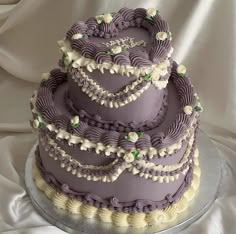 a three tiered cake with purple icing and white flowers on top is sitting on a silver platter