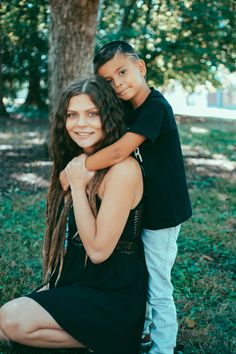 a woman holding a young boy while sitting on the ground in front of a tree