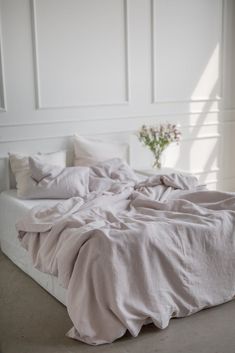 an unmade bed in a white room with flowers on the nightstand and wall behind it