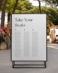 a sign that says take your seats in front of some trees and flowers on the sidewalk