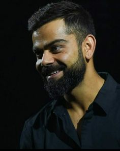 a man with a beard and black shirt smiling at the camera while standing in front of a dark background