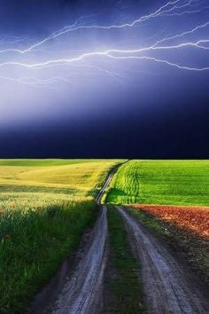 a dirt road in the middle of a field under a stormy sky with lightning above it