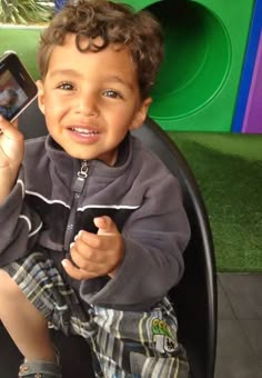 a young boy sitting in a chair holding a cell phone and smiling at the camera