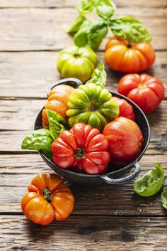 several different colored tomatoes in a metal bowl on a wooden table with basil leaves around them