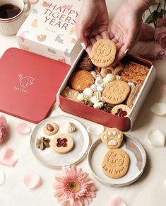 two hands grabbing cookies out of a red box on a table with pink flowers and petals