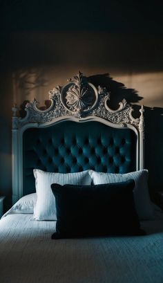 a bed with a blue headboard and white pillows in a dark room, lit by light coming from the window