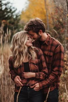 a man and woman standing together in tall grass with their arms around each other, looking into each others eyes