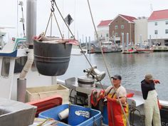 two men are standing on the deck of a boat near other boats and buildings in the water