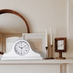 a white clock sitting on top of a mantle next to candles and framed pictures in front of a mirror