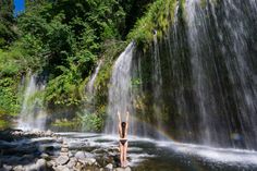 a woman standing in front of a waterfall with a rainbow above her head and arms up