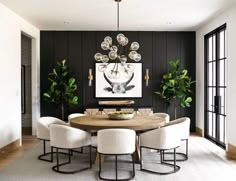 a dining room table surrounded by white chairs and potted plants in front of a black wall