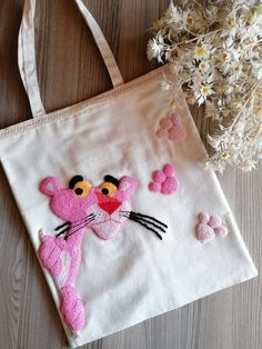 a pink cat embroidered onto a white canvas bag next to some daisies and flowers