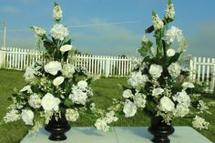 two black vases filled with white flowers sitting on top of a cement slab in the grass