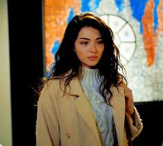 a woman with long hair wearing a tan coat and white turtle neck sweater standing in front of a painting