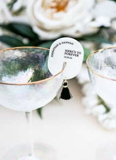 two wine glasses sitting next to each other on top of a white table with flowers in the background