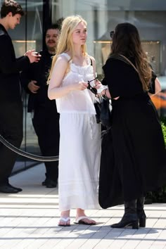 two women in white dresses talking to each other on the sidewalk with people standing around