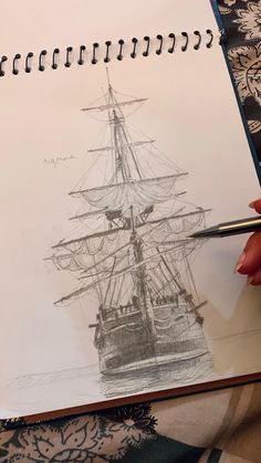 a drawing of a ship on paper next to a person's hand holding a pen