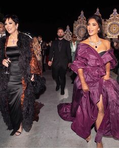 two women in purple dresses walking next to each other at a formal event with people dressed in black