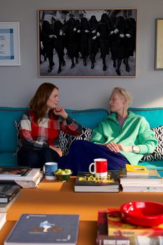 two women sitting on a couch talking to each other in front of pictures and books