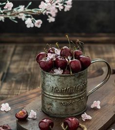 cherries are in a metal cup on a wooden board with cherry blossoms around it