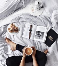 a person laying in bed reading a book and drinking coffee