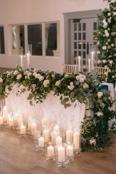 candles are lined up on the table with flowers and greenery around them, along with white roses