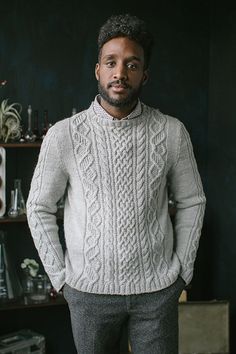 a man standing in front of a black wall wearing a white sweater and gray pants