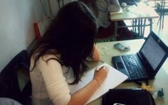 a woman sitting at a desk writing on a piece of paper with a laptop in front of her