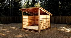 a small wooden shelter in the middle of a gravel area with trees and fence behind it