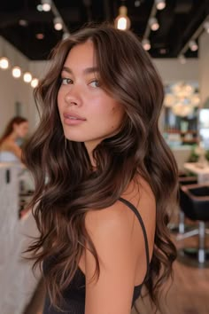 Woman with long, wavy brown hair in a well-lit salon. Rich Hair Color Ideas For Brunettes, Chocolate Brown Dark Hair, Brown Hair Colors For Asians, Dark Brown Hair Balayage Chocolate, Long Hair Chocolate Brown, Hair Gloss For Brunettes, Hair Color For Medium Dark Skin Tones, Glowy Brown Hair, Rich Milk Chocolate Brown Hair