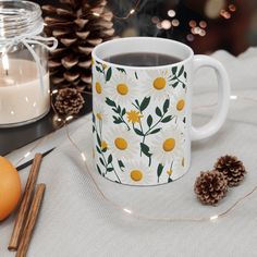 a coffee mug with daisies on it next to some cinnamons and pine cones