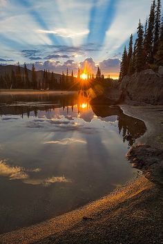 the sun is setting over a lake with rocks and trees in the foreground, while clouds are reflected in the water