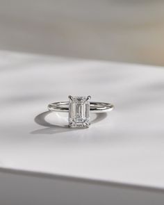 an emerald cut diamond ring sitting on top of a white surface with shadow from the background