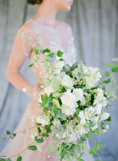 a woman in a wedding dress holding a bouquet of white flowers and greenery with her hands behind her back